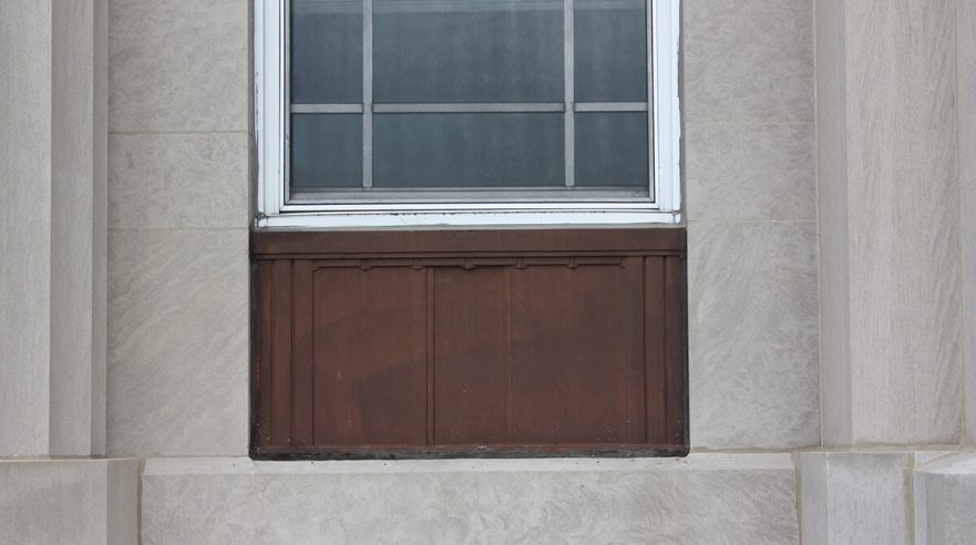 Small metal panels that are beneath the first floor windows on the west façade of City Hall, note they have minimal decorations, circa 2022.